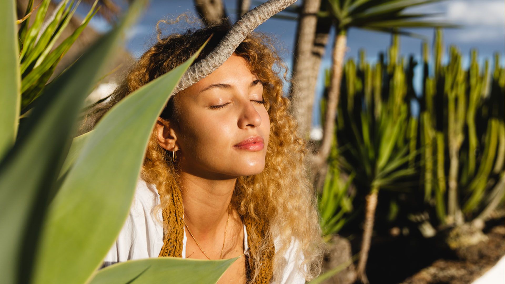 Woman looking serene and relaxed with her eyes closed sits in a sunny botanic garden surrounded by large, leafy green plants.