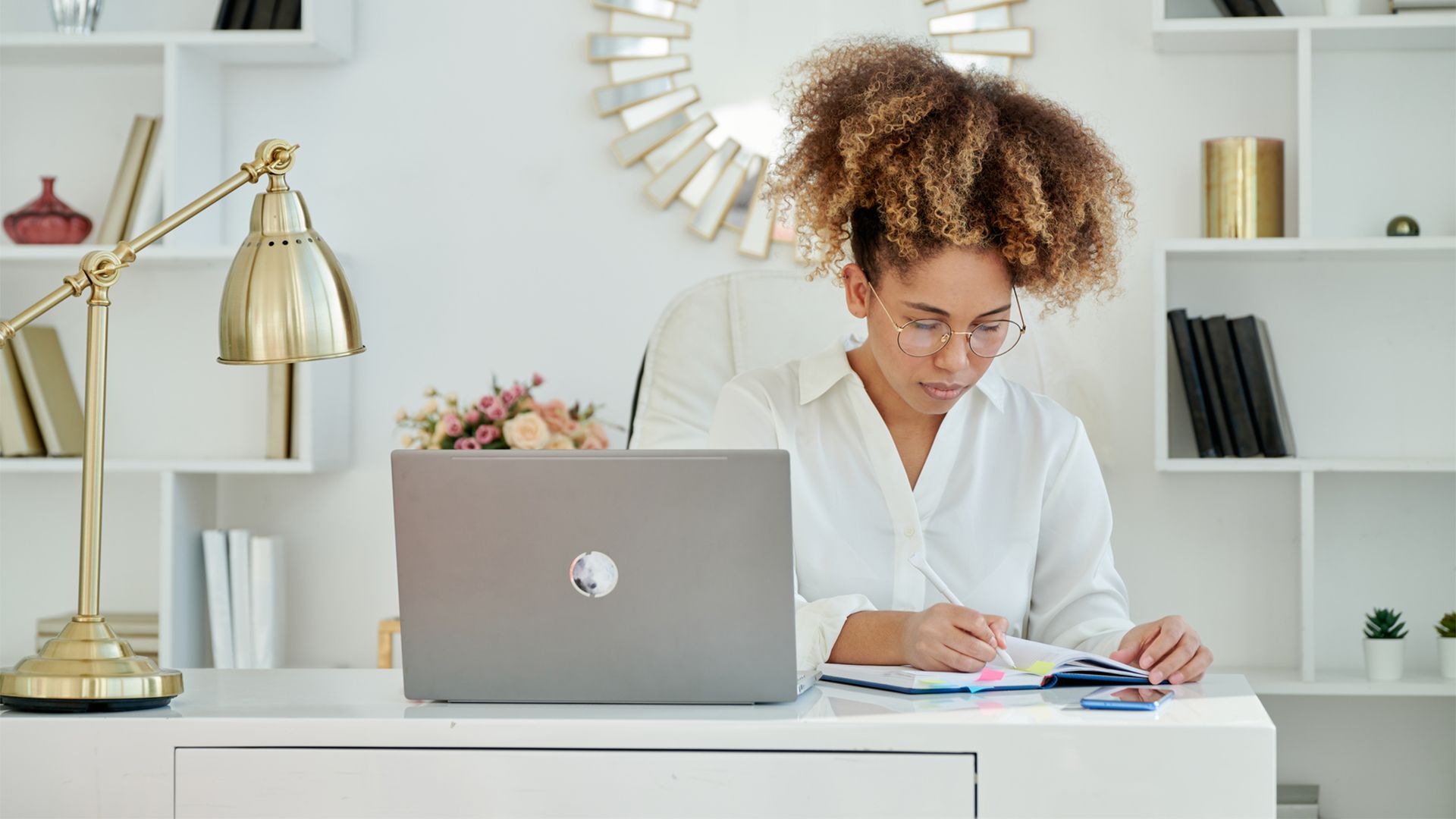 A young Black woman wearing a white button-down shirt and glasses sits at a desk writing in a journal while her laptop is open next to her.