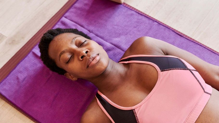 Black Woman with short hair and wearing a sports bra lays on a yoga mat with her eyes closed.