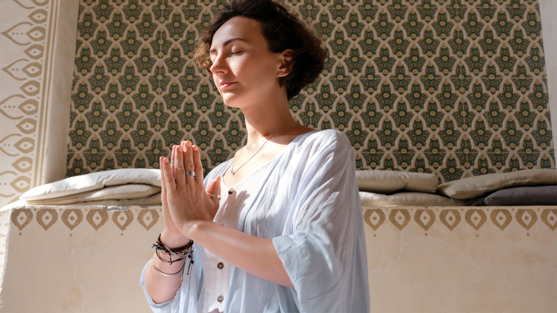 Low angle photograph of a white woman with closed eyes gesturing namaste and meditating against the wall with arabesque ornaments.