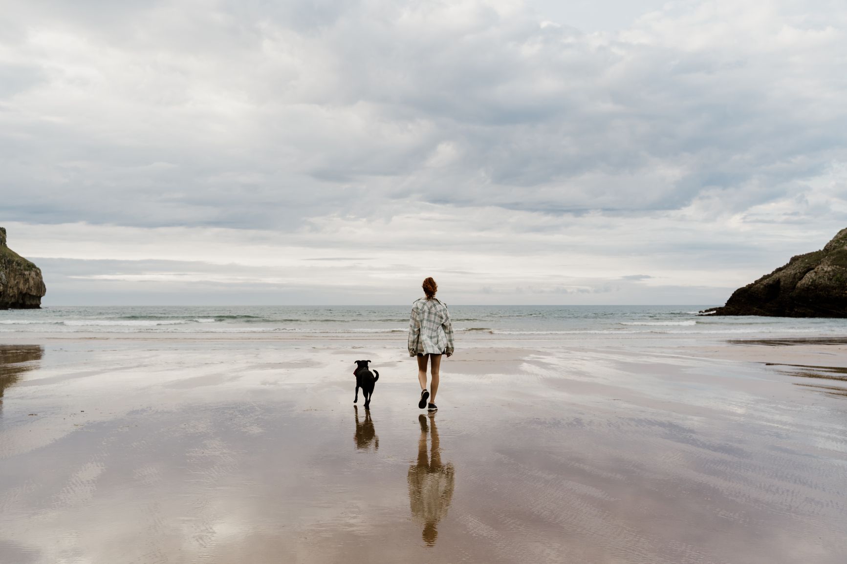 Woman and dog walk along a quiet beach during the day 