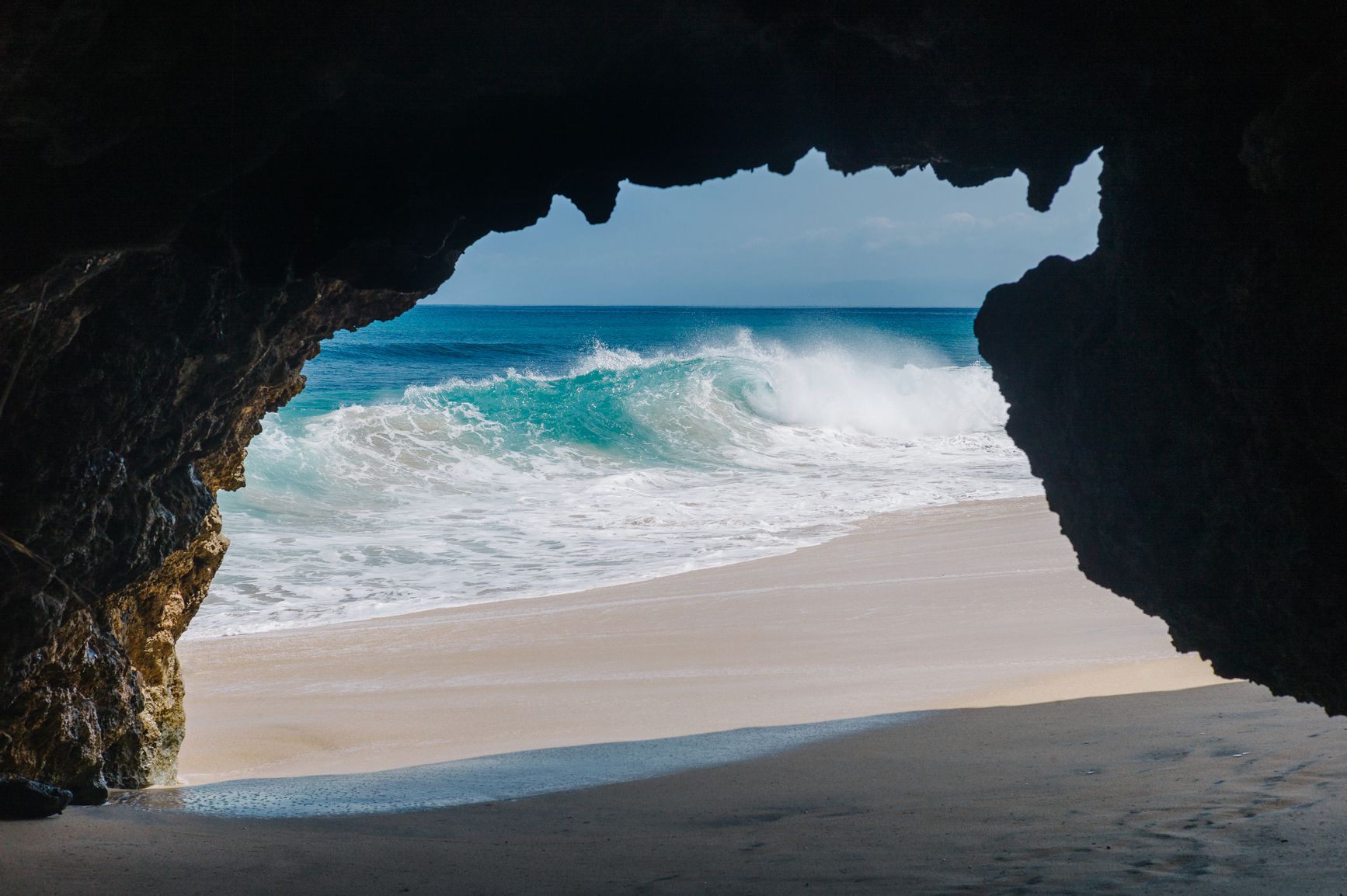 View from inside a cave facing out to the blue ocean waves and a sandy shore