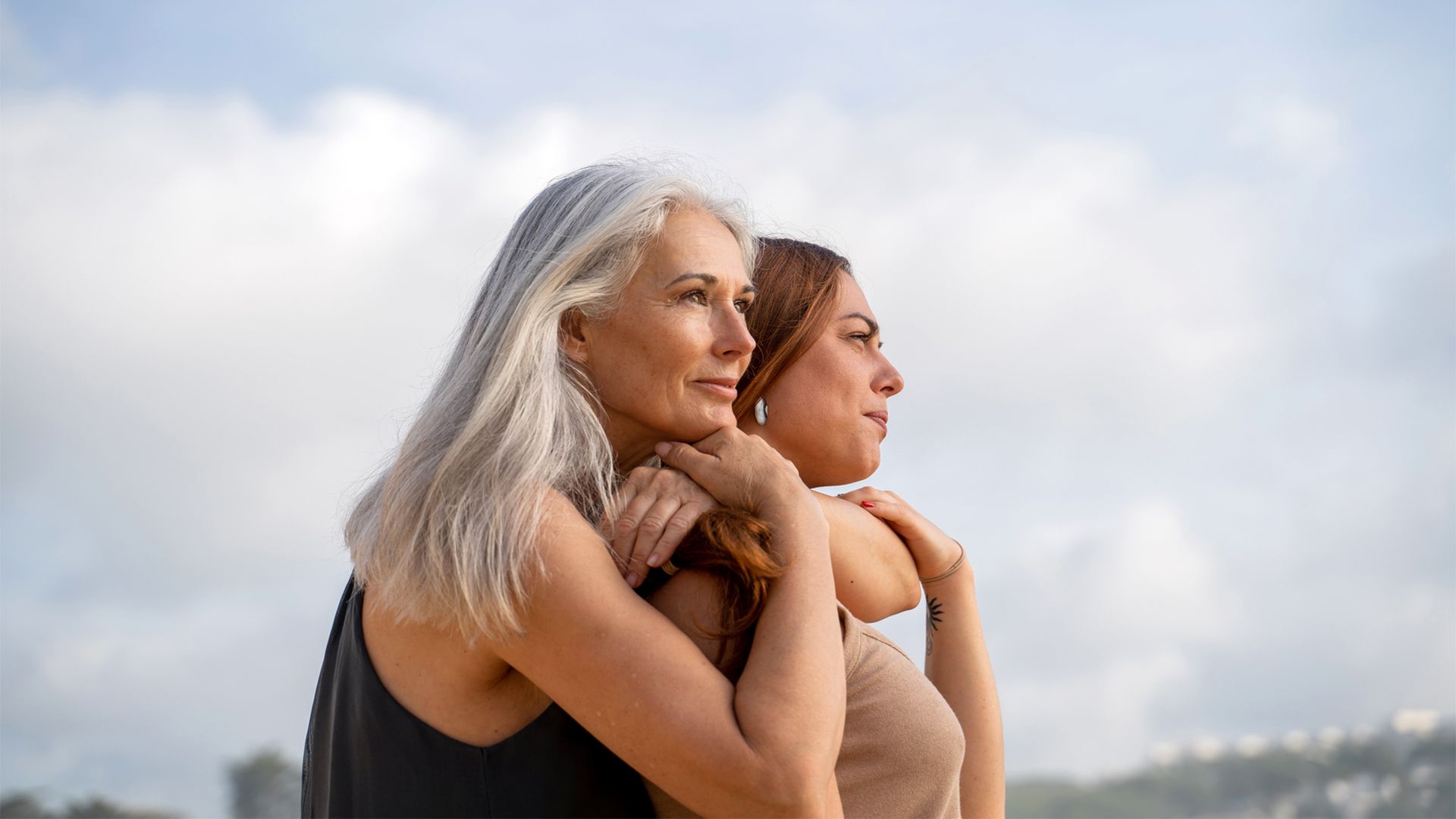 Mother and adult-aged daughter embrace while standing on the beach.