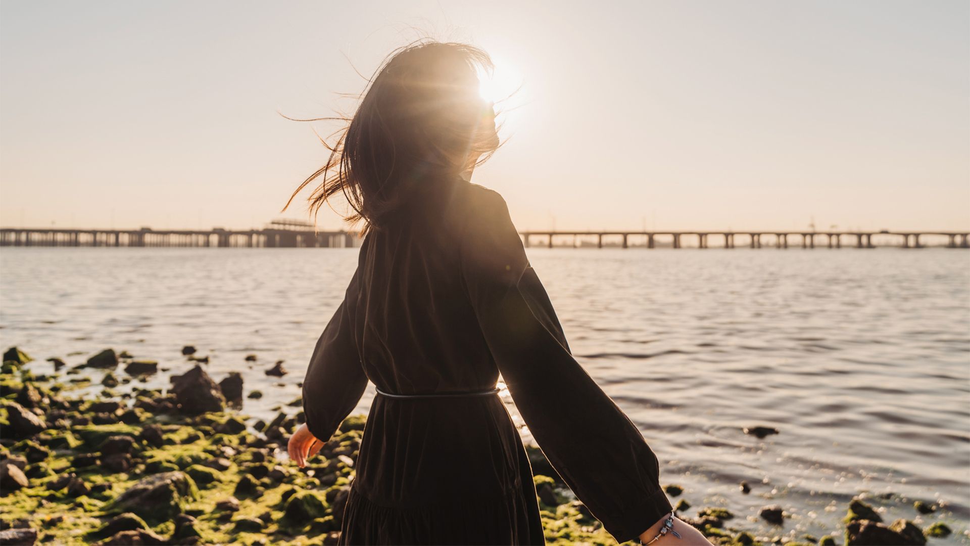 A portrait of a woman wearing a black dress with her arms stretched out at her sides being backlit by the sun on the beach.