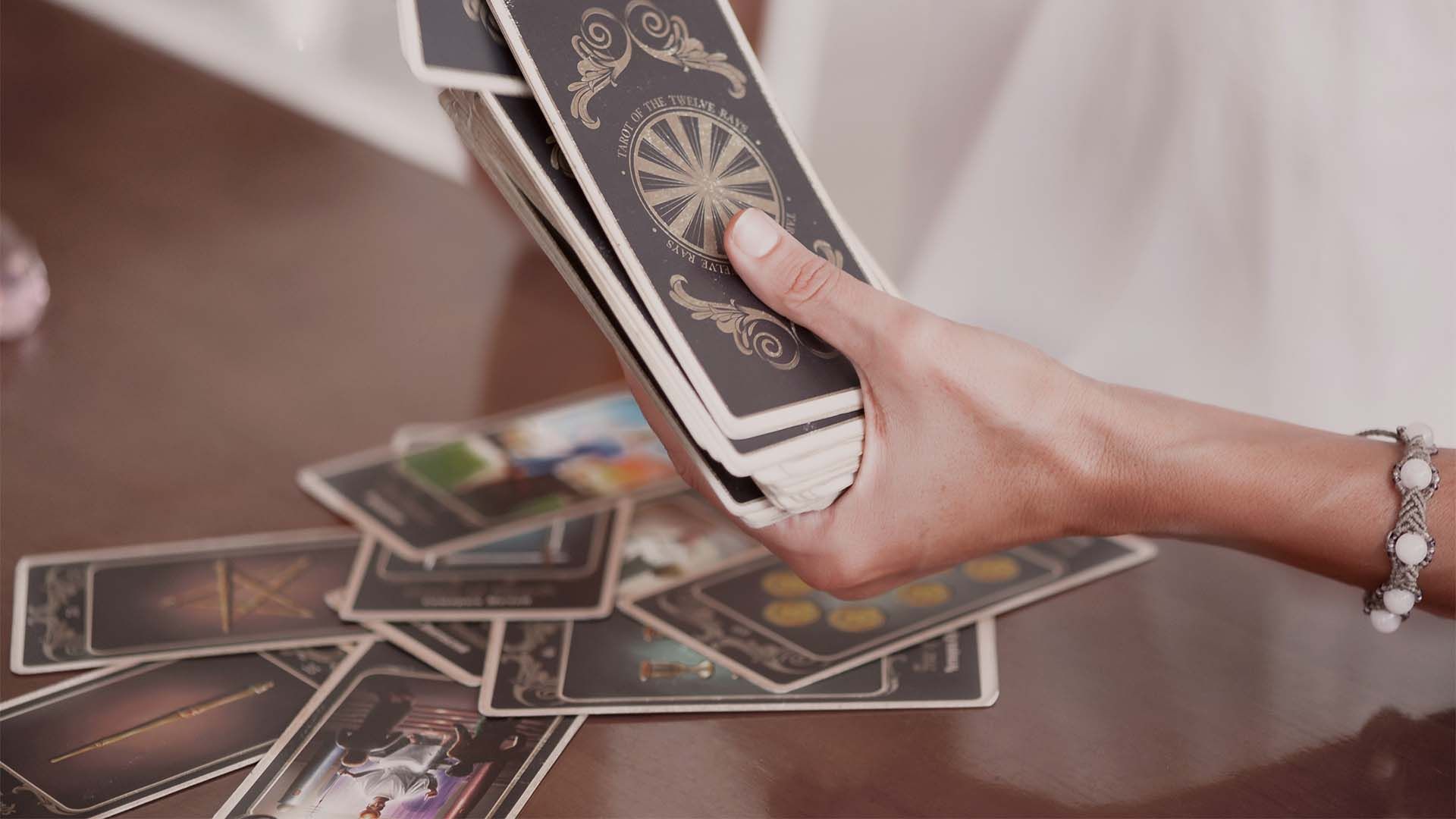 A hand with a bracelet holds a deck of Tarot cards, with more spread out on a table beside her.