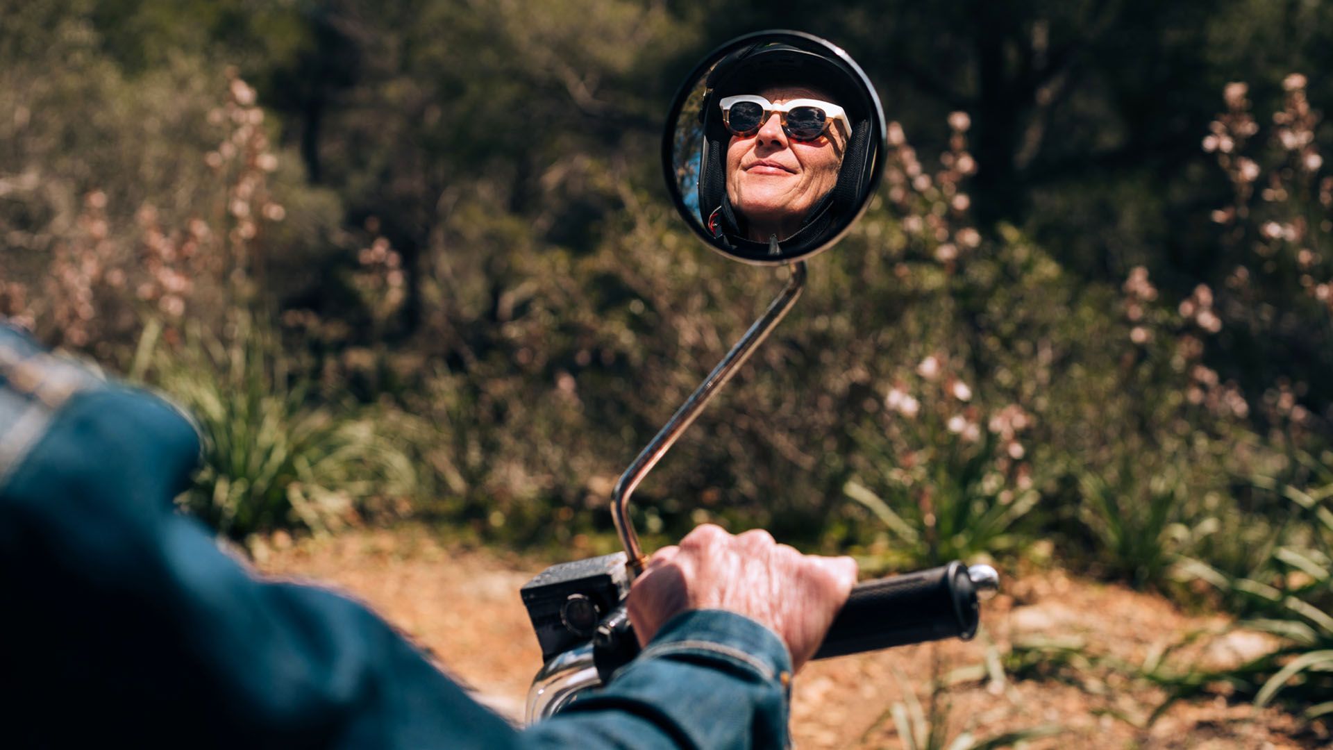 Senior-aged woman wearing sunglasses rides on a motorcycle, her smiling reflection shows in the motorcycle’s mirror.