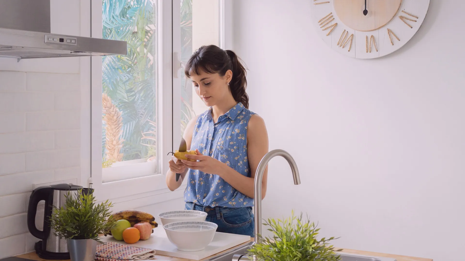 Young white woman standing in a sunny, bright kitchen prepares a meal in front of two bowls and a variety of fresh fruits.