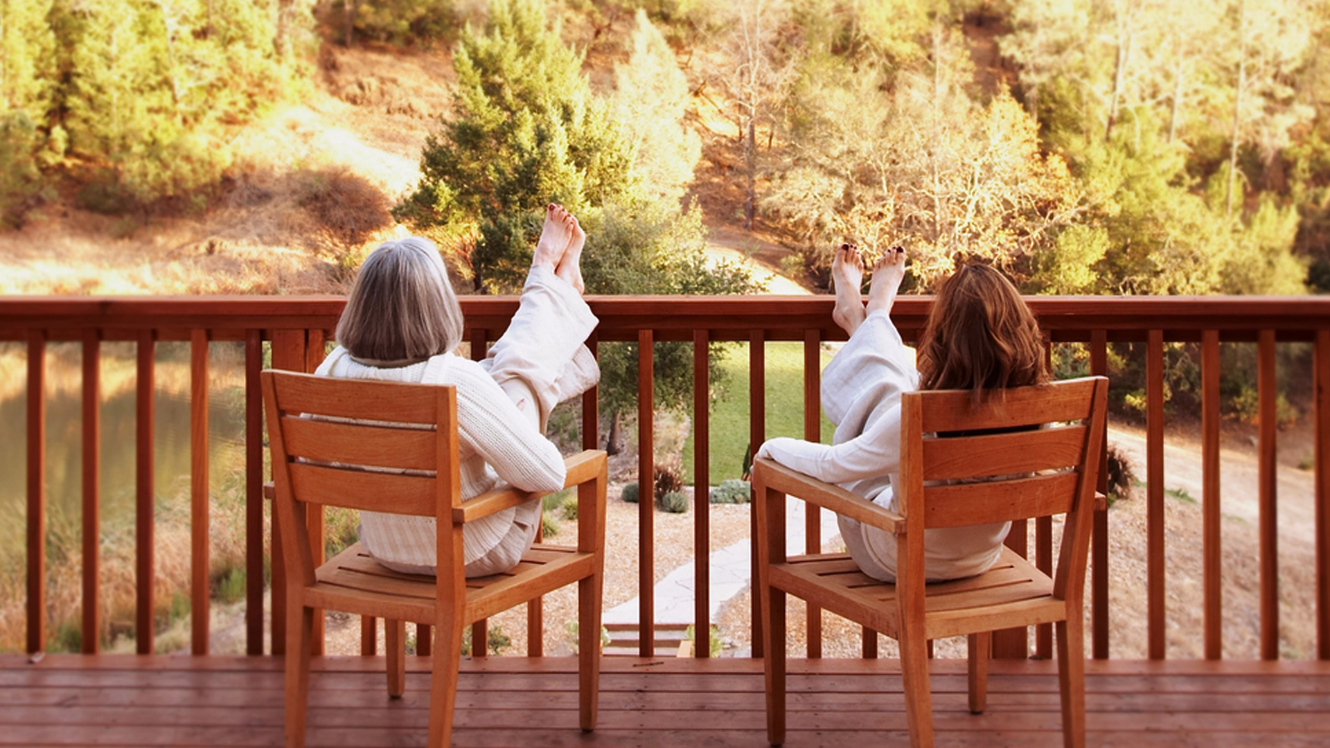 Two women, both wearing light-colored clothing, sit on an outdoor wooden deck with their feet resting in front of them as they look at a peaceful nature setting with trees and a lake. .