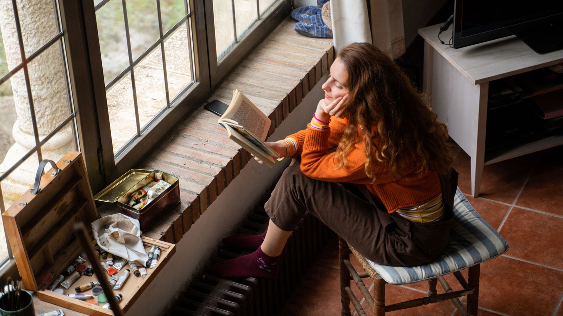 White woman with long brown hair sits on a chair by a windo reading a book.