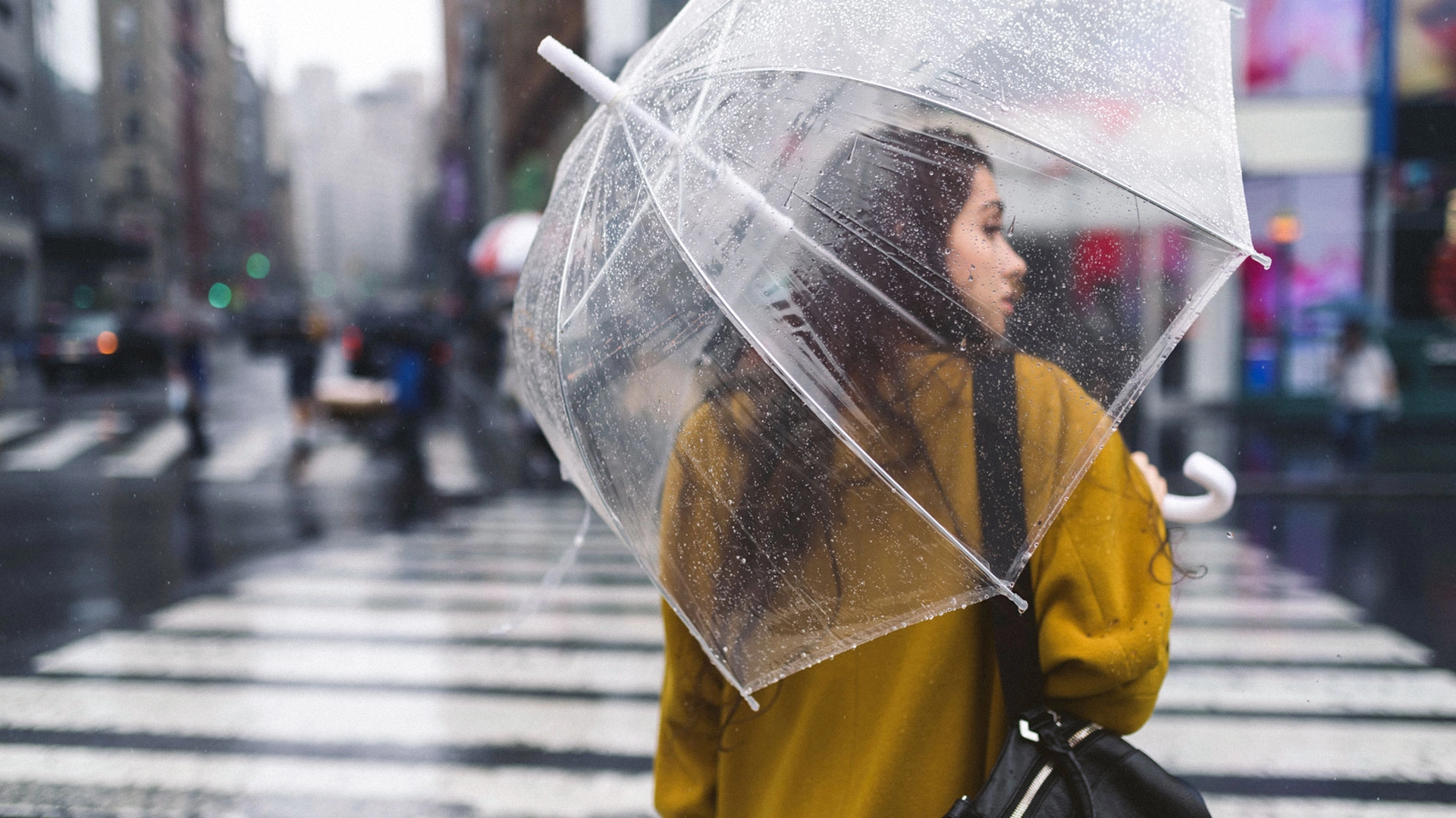 Woman wearing mustard yellow coat walking in the rain holding an umbrella while crossing a city street.