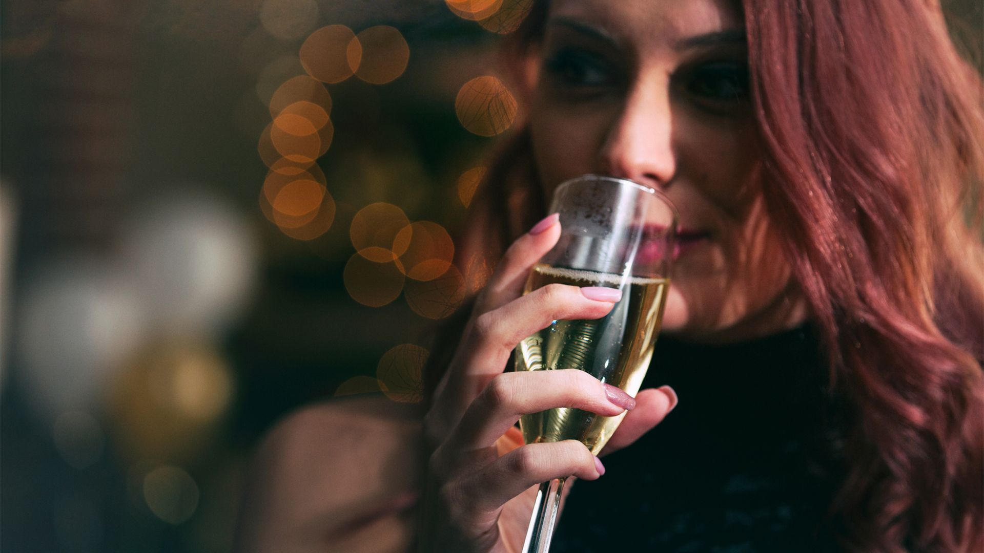 A young white woman with long red hair smiles slightly as she drinks champagne from a champagne flute.