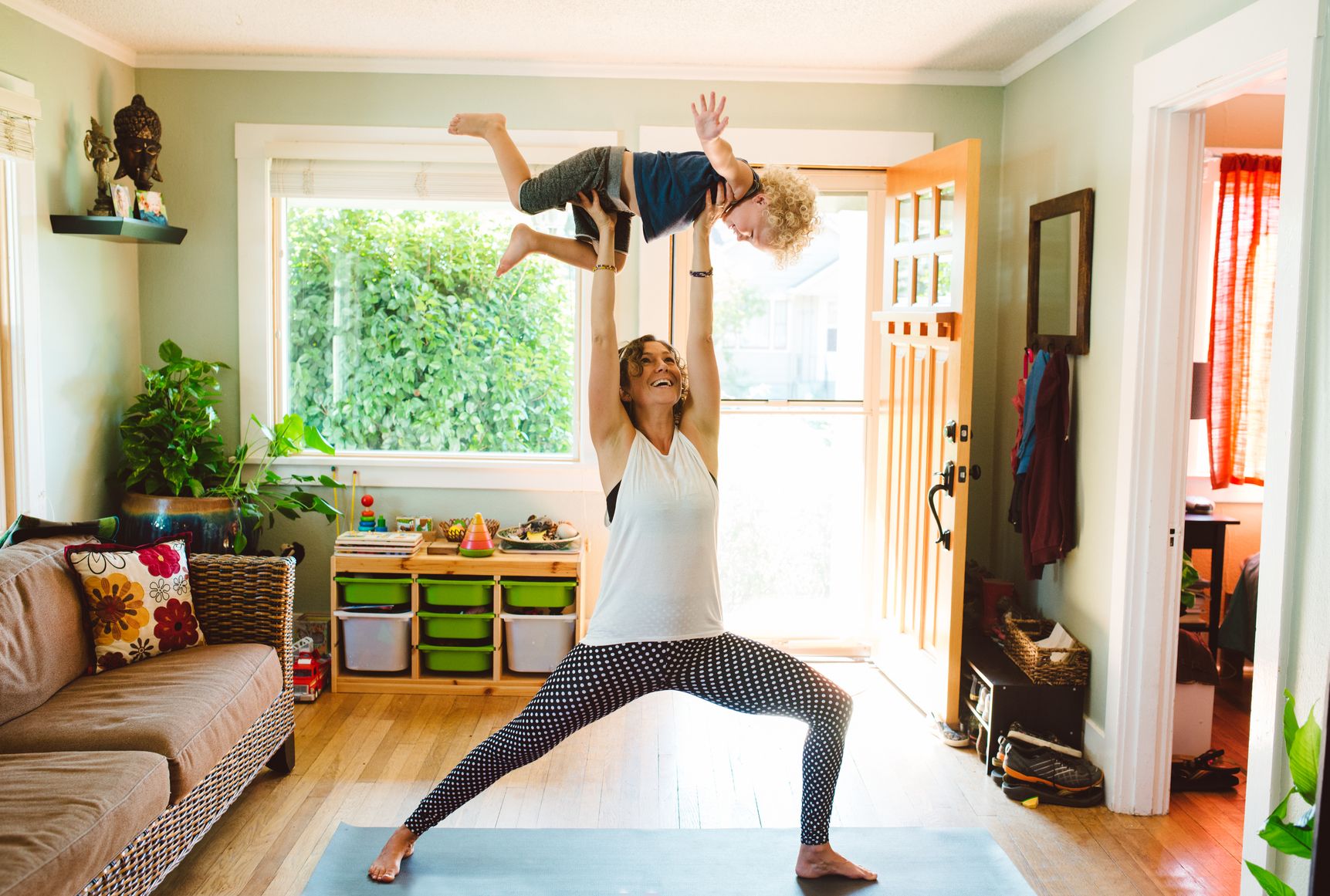 Laughing mom in exercise clothing and standing on a yoga mat holds her toddler above her head as they exercise together