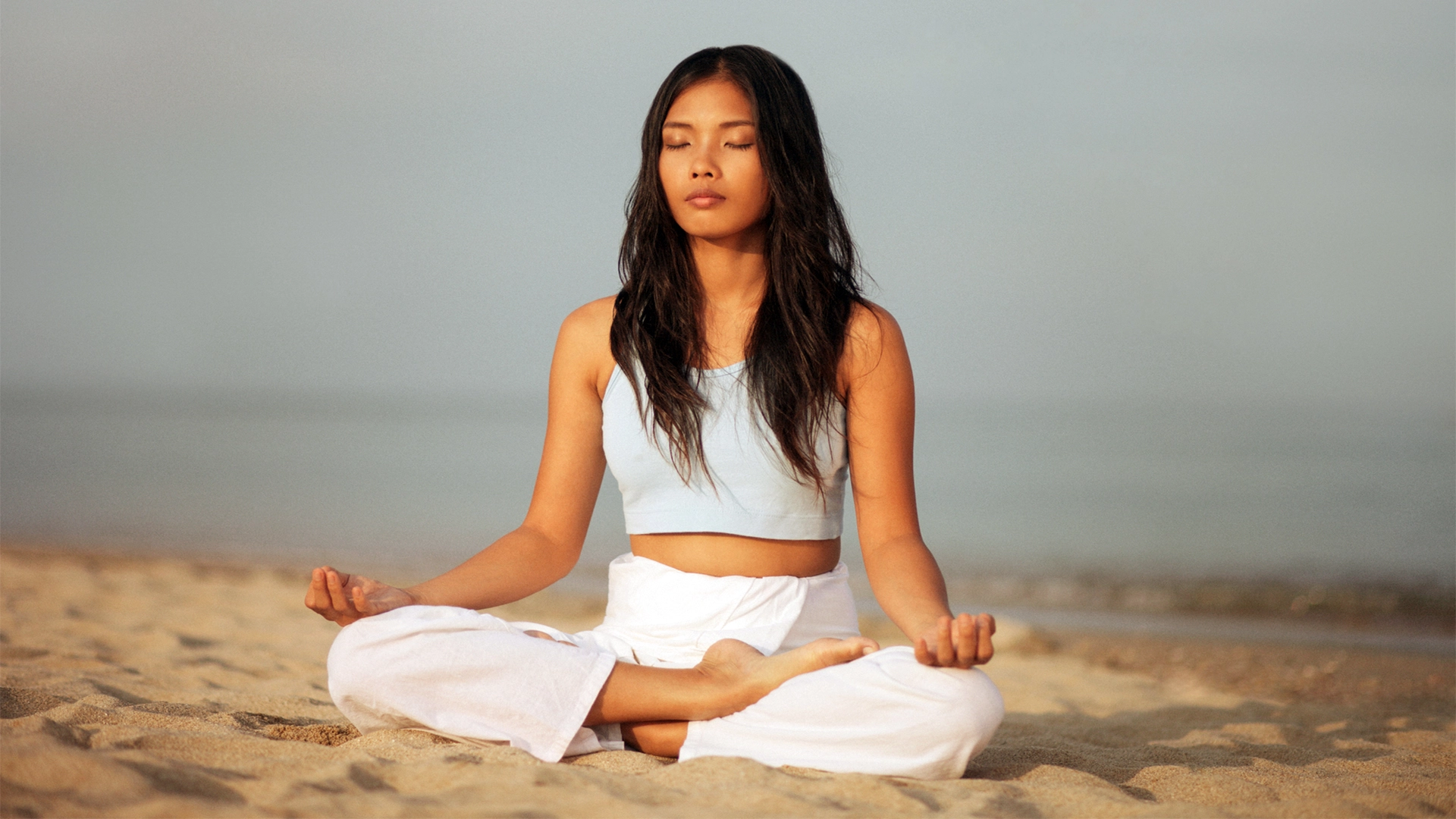 Young Asian woman wearing white pants and a white tank top sits on the beach in a cross-legged pose, her eyes closed, meditating.