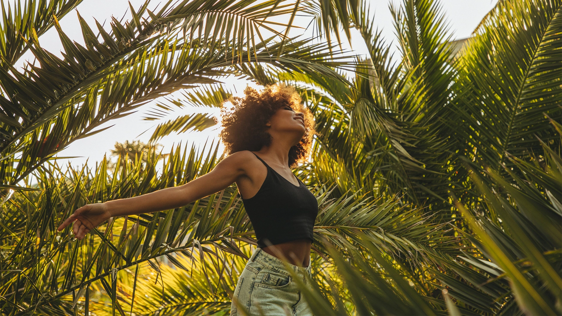 A young woman with an afro who is wearing a blank tank top and jeans stands smiling underneath palm leaves on a sunny day.