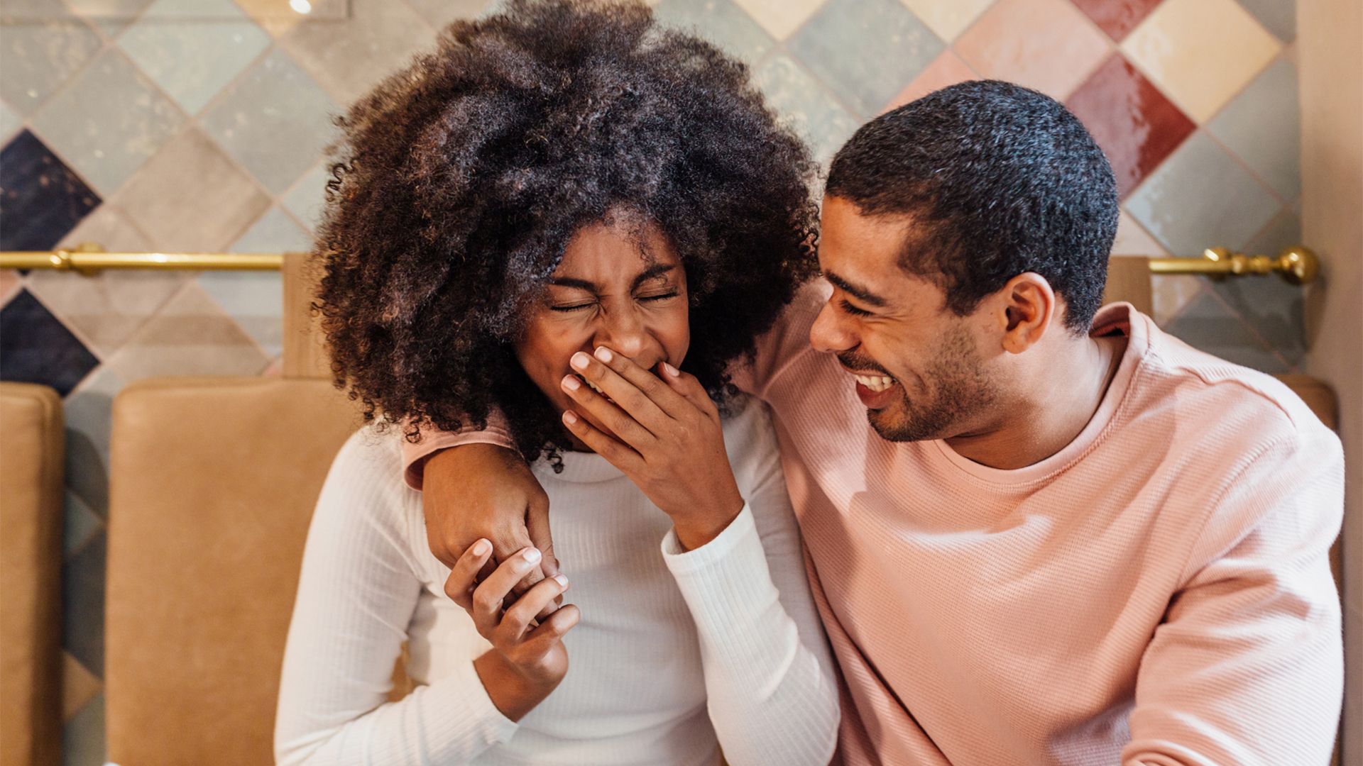 A happy couple photograph features a smiling Black man wearing a pink sweatshirt and a laughing Black woman in a white long-sleeve t-shirt sitting close together in a cafe, his arm around her shoulder, as she laughs and covers her mouth at something he says.