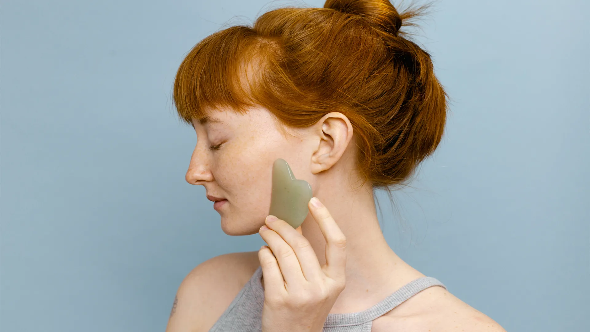 Peaceful looking younger white woman with red hair in a bun holds a jade Gua Sha tool near her face.