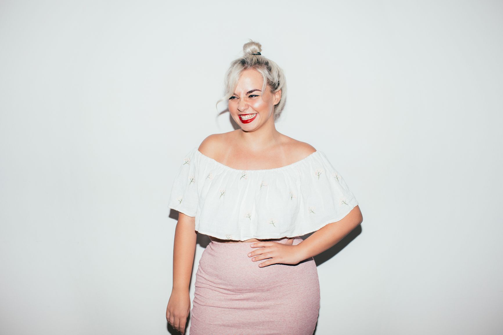 Woman wearing white off-the-shoulder top and pink skirt. Smiles against a white wall. 