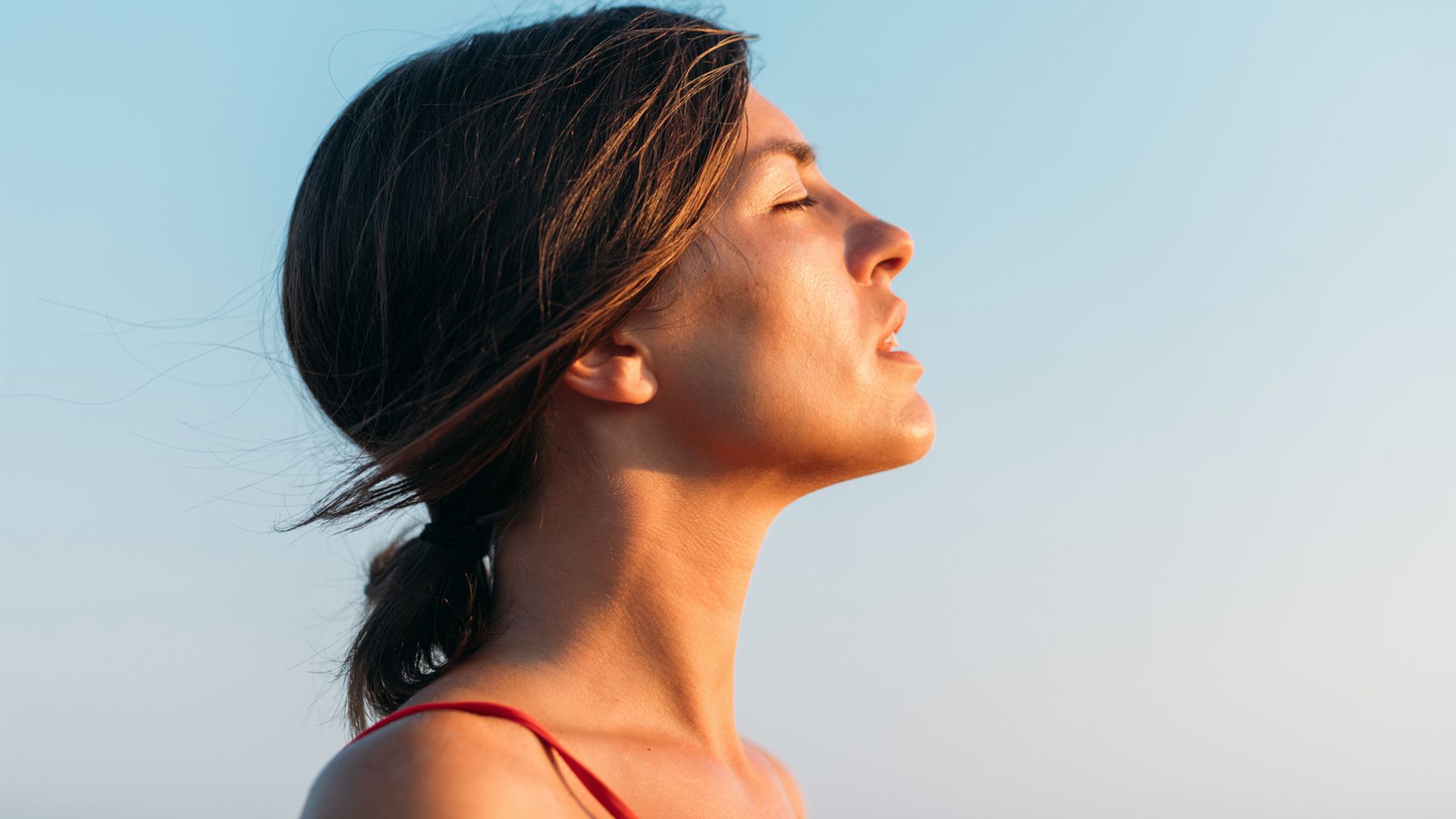 A young white woman with brown hair pulled back into a ponytail stands outdoors on a sunny day, with her eyes closed.