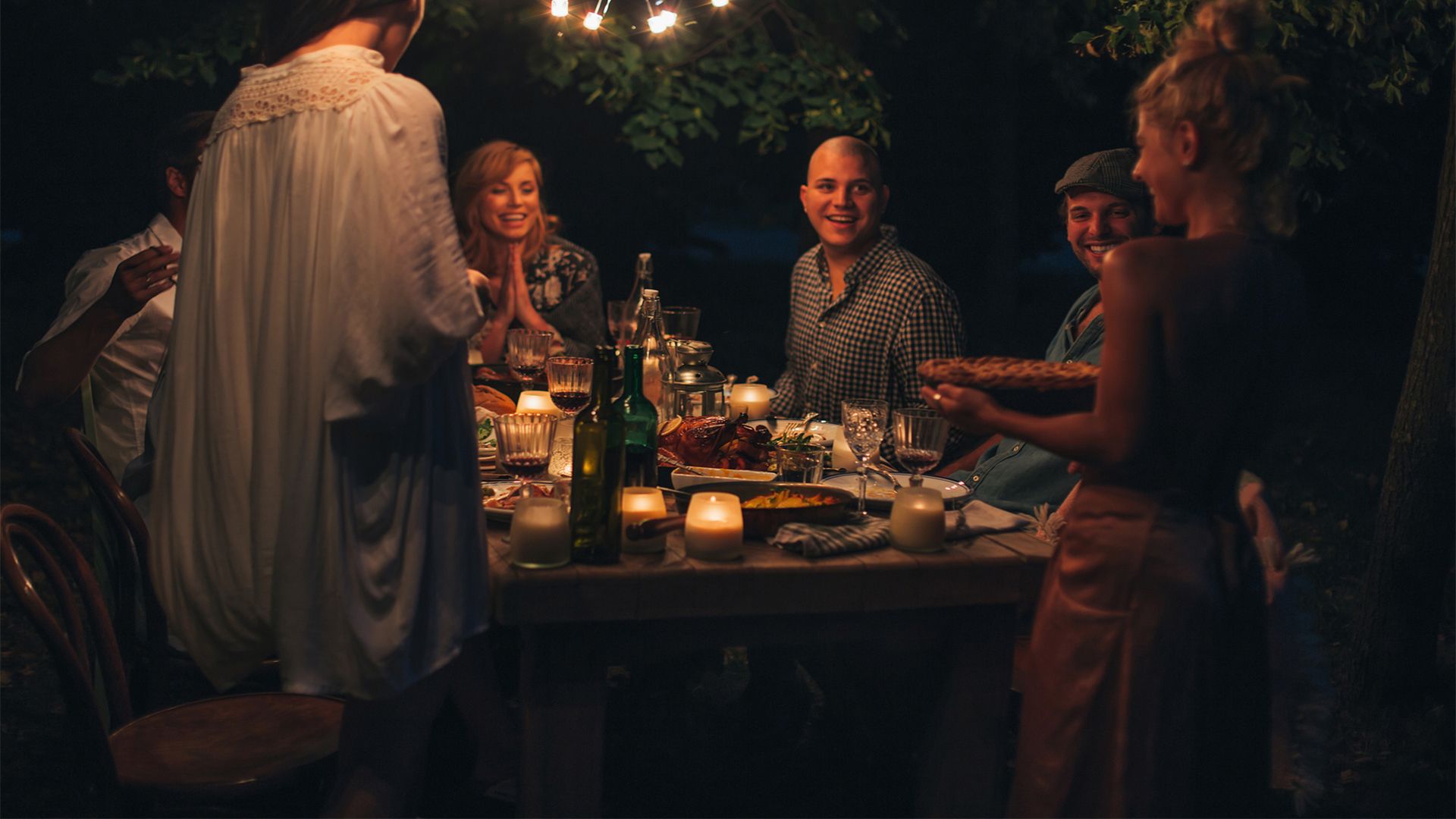 A group of six friends looking happy and smiling gather around a table that has dishes, wine bottles, and candles, to share a meal outdoors at night under twinkling lights.